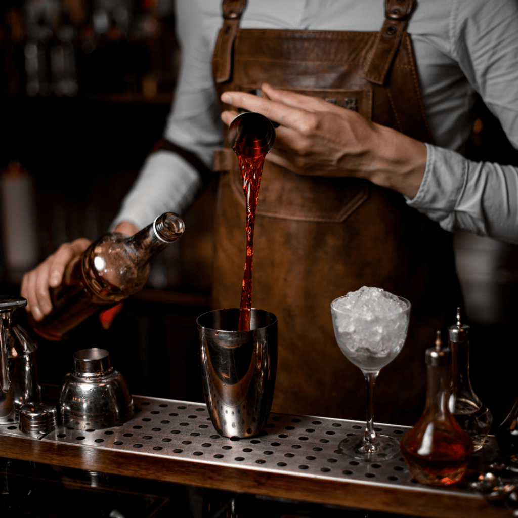 A Thirst bartender creates a bold cocktail moment, expertly pouring a deep red spirit into a shaker. Dressed in a leather apron, they work behind a sleek bar setup, with a chilled coupe glass filled with ice ready for the final touch.