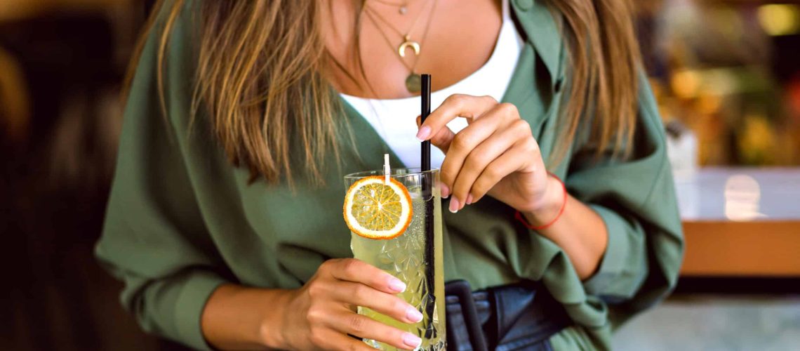 Close up cropped image of woman holding sweet tasty cocktail lemonade, stylish trendy hipster outfit and elegant jewelry, toned colors, bar atmosphere.